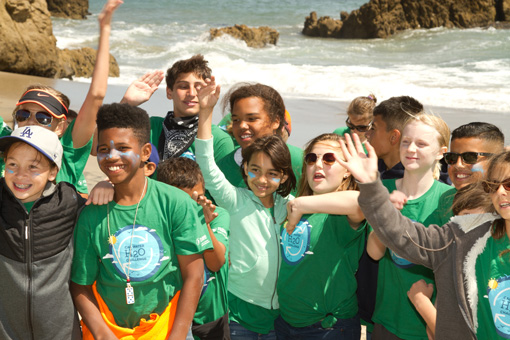 Photograph of smiling 5th grade students with the Pacific ocean at their backs.  A few children are raising their hands.