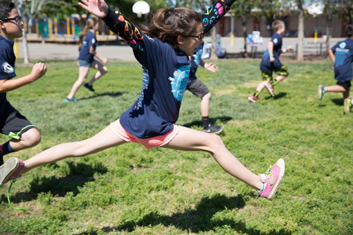 Photograph of a group of 6th grade children happily running, in the forground a girl is leaping, both feet off the ground, one shoe untied.