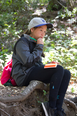 Photograph of a 6th grade girl sitting, thinking, on a log, holding a pencil with a pink notebook resting on her lap.